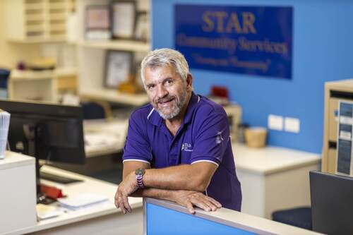 Jeff smiling in his purple uniform top leaning on an office wall