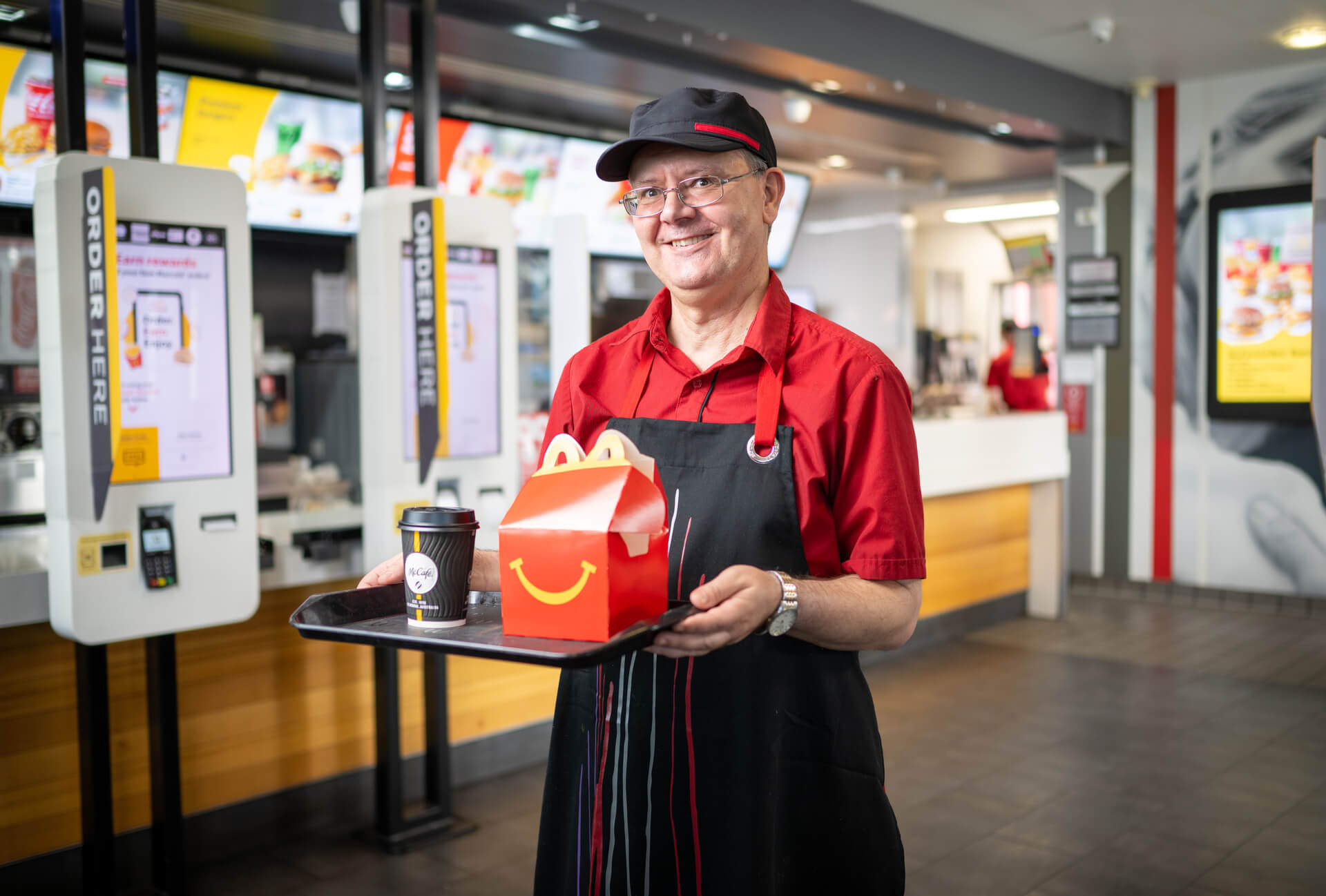 A man in a McDonald's restaurant smiles as he holds a tray with a drink and McHappy Meal