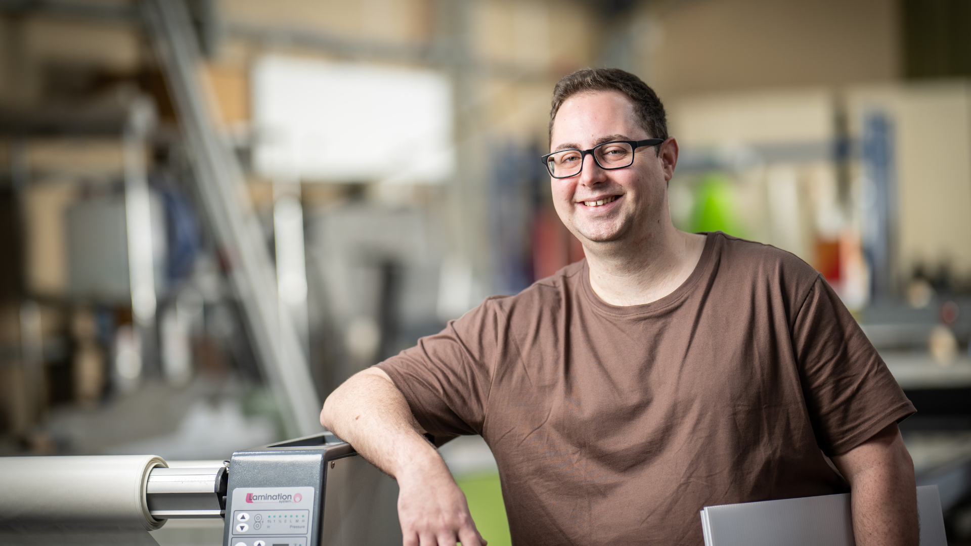 Sean in a brown tshirt leaning on a machine in a workshop