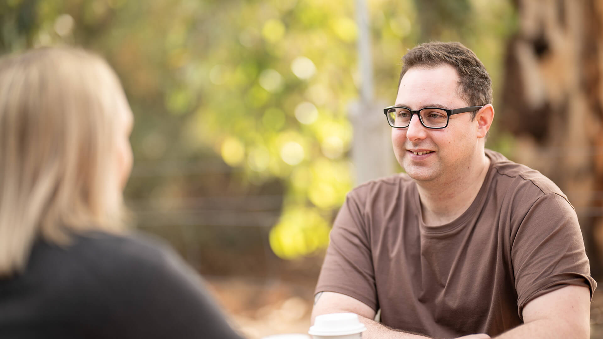 Sean lives with Crohn's disease, smiles in a brown tshirt sat outside 