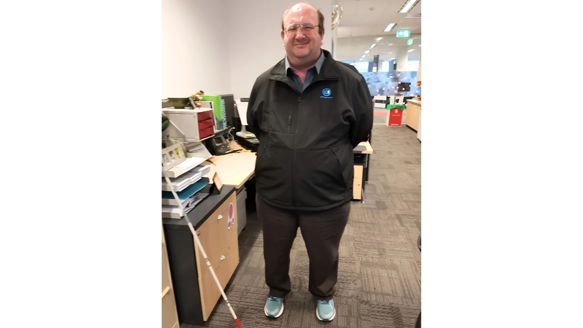 A man standing in an office with a white cane leaning on a desk